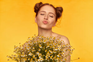 Mujer pelirroja con moños y ojos cerrados lanzando un beso sobre fondo amarillo con ramo de manzanilla