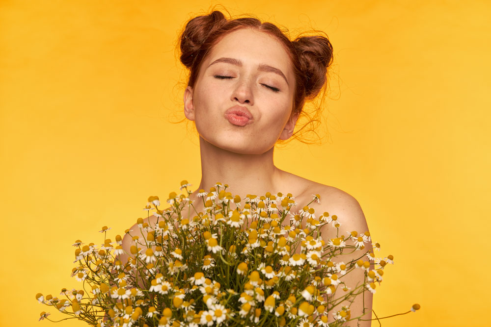 Mujer pelirroja con moños y ojos cerrados lanzando un beso sobre fondo amarillo con ramo de manzanilla