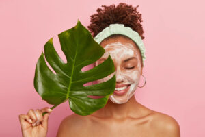Mujer sonriente con espuma limpiadora facial y diadema sostiene una hoja de monstera sobre fondo rosa