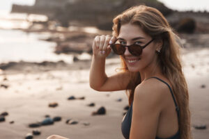 Mujer sonriente en la playa al atardecer bajando sus gafas de sol junto a rocas y arena