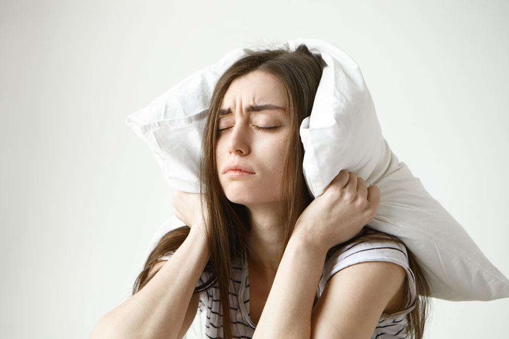 Mujer con los ojos cerrados y gesto de agotamiento tapándose los oídos con una almohada