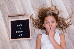 Niña con cara de sorpresa tumbada junto a un cartel con el texto Back to School y un lápiz