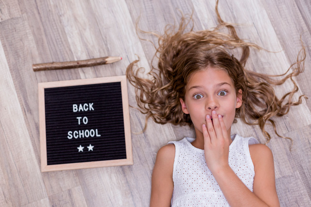Niña con cara de sorpresa tumbada junto a un cartel con el texto Back to School y un lápiz