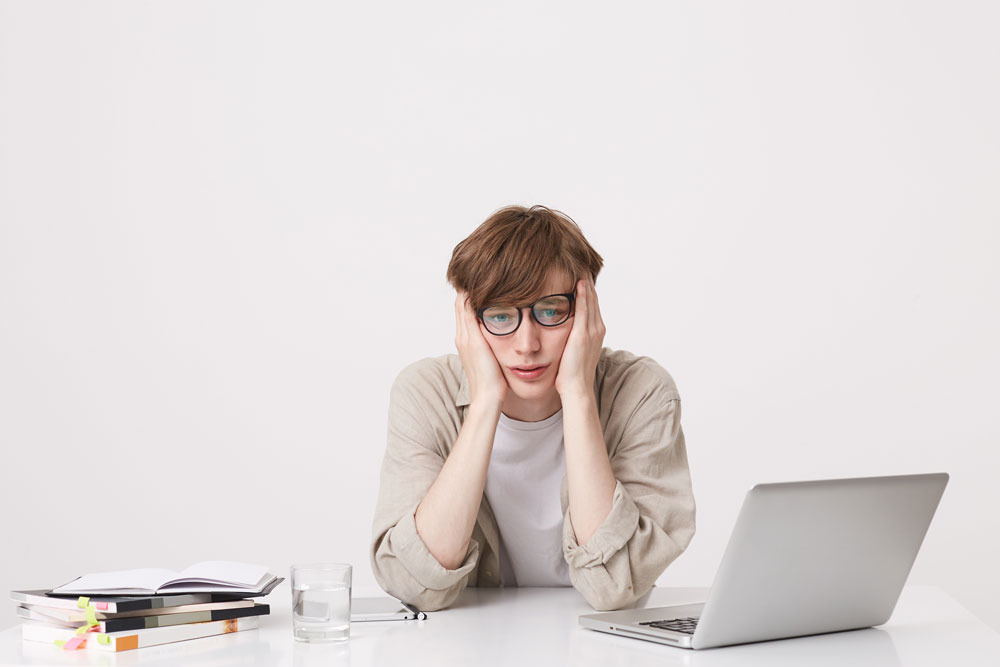 Joven con gafas y aspecto cansado apoya la cabeza en las manos frente a un portátil en su escritorio