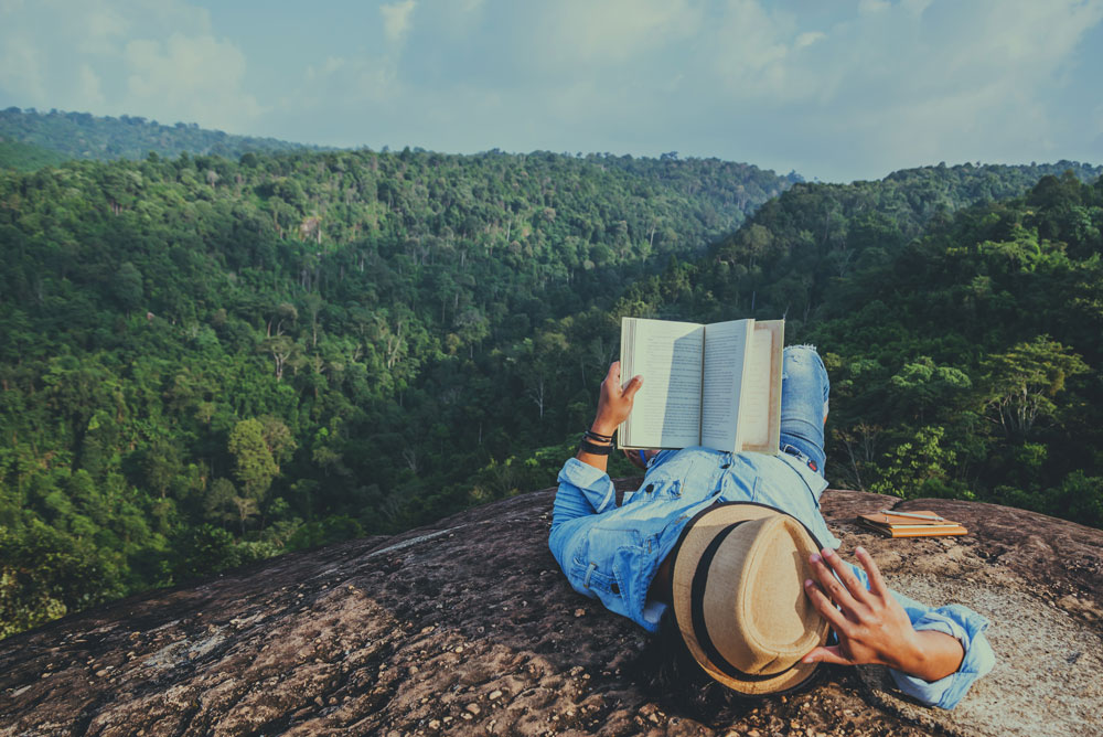 Persona tumbada en una roca leyendo un libro frente a un valle montañoso lleno de bosque verde