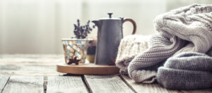 Cafetera gris, jarrón con lavanda y jerséis de punto sobre mesa de madera junto a una ventana