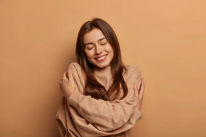 Mujer joven sonriente con jersey beige se abraza a sí misma con los ojos cerrados