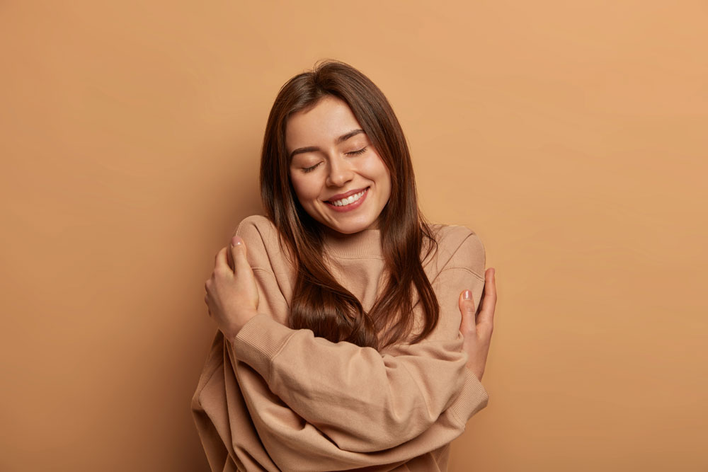 Mujer joven sonriente con jersey beige se abraza a sí misma con los ojos cerrados