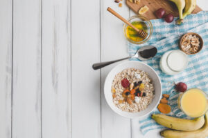 Bol de muesli con frutos secos, miel, leche, plátanos y zumo sobre mesa de madera blanca