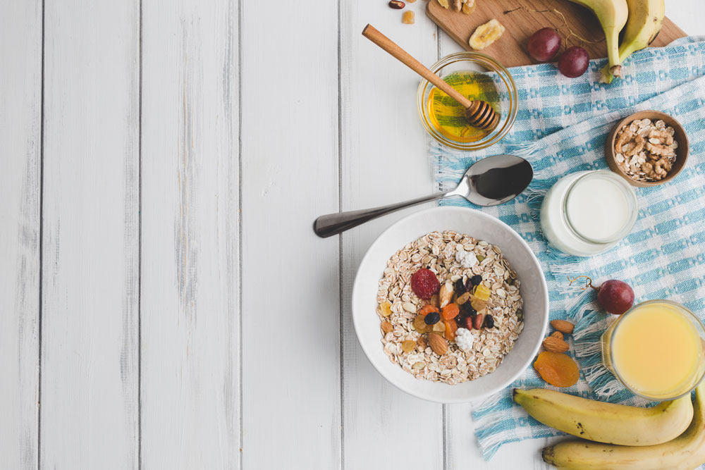 Bol de muesli con frutos secos, miel, leche, plátanos y zumo sobre mesa de madera blanca