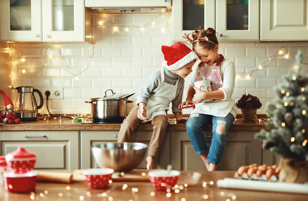 Dos niños con gorro de Papá Noel sentados en la encimera de una cocina decorada con luces de Navidad