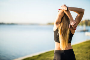 Mujer con top deportivo estira el brazo tras la nuca junto a un lago al atardecer