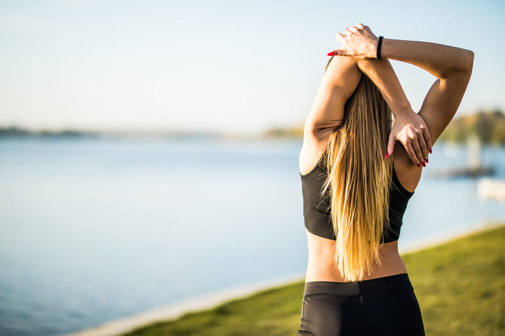 Mujer con top deportivo estira el brazo tras la nuca junto a un lago al atardecer