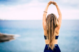 Mujer de espaldas estirando los brazos hacia el cielo frente al mar al aire libre