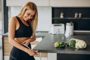 Mujer sonriente en ropa deportiva midiendo su cintura junto a frutas y verduras en la cocina