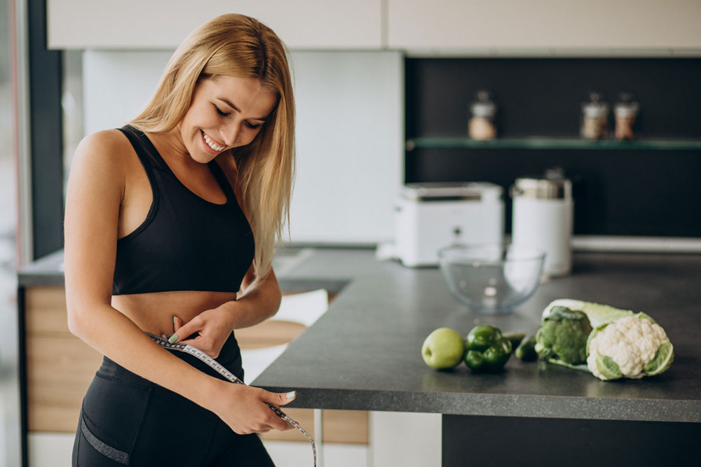Mujer sonriente en ropa deportiva midiendo su cintura junto a frutas y verduras en la cocina