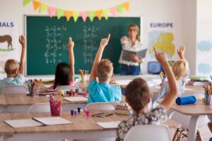 Niños en el aula levantando la mano ante la pizarra con sumas mientras la maestra da clase