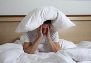 Hombre en la cama con una almohada sobre la cabeza y gesto de cansancio por jet lag