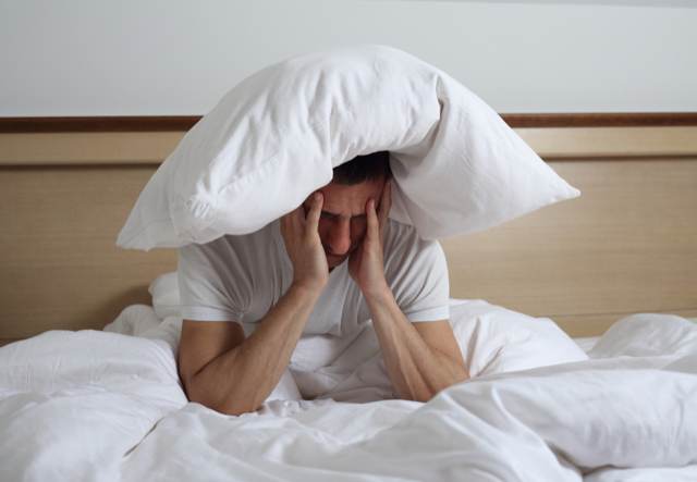 Hombre en la cama con una almohada sobre la cabeza y gesto de cansancio por jet lag