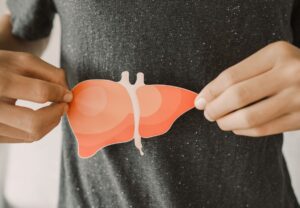Manos sostienen recorte de papel con forma de hígado sobre camiseta gris a la altura del abdomen