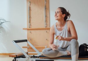 Mujer sonriente sentada en un reformer de pilates sostiene una botella de agua tras entrenar