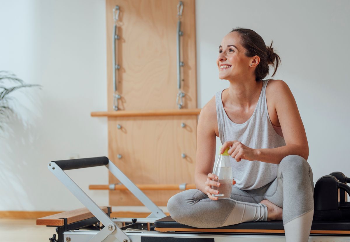 Mujer sonriente sentada en un reformer de pilates sostiene una botella de agua tras entrenar