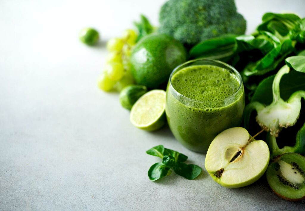 Smoothie verde rodeado de frutas y verduras frescas como manzana, kiwi, brócoli y espinacas