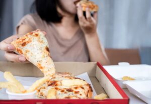 Mujer levanta una porción de pizza con queso derretido junto a patatas fritas en una caja