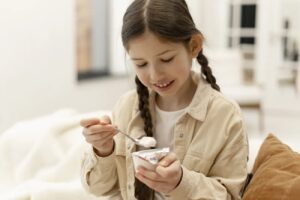 Niña con trenzas sonriendo mientras come yogur con cuchara, cuidando su flora intestinal