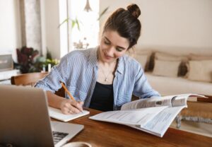 Mujer joven sonriente estudiando y tomando notas con un libro y un portátil sobre la mesa