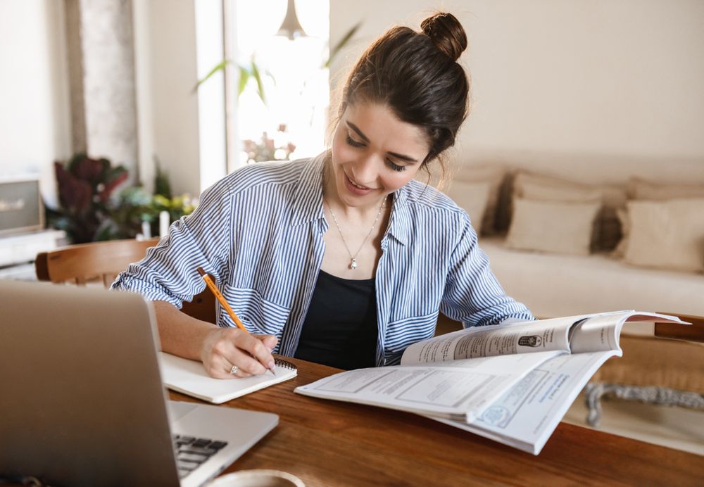 Mujer joven sonriente estudiando y tomando notas con un libro y un portátil sobre la mesa